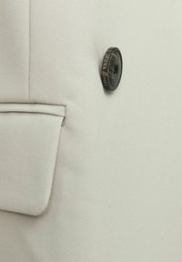 Chaqueta beige con una textura de tela suave, que cuenta con un bolsillo lateral y un botón negro redondo con la etiqueta "Jack & Jones."