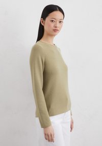 Olive green long-sleeve sweater with a round neckline and textured fabric, paired with white pants. Simple design, fitted silhouette.