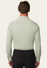 Lichtgroene blouse met lange mouwen en een klassieke kraag, met knoopmanchetten, gemaakt van een gladde stof, gezien van achteren. Zwarte broek zichtbaar.