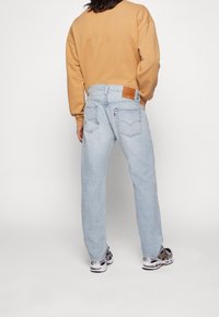 Person wearing light blue denim jeans, tan sweatshirt, and black and white sneakers, standing facing away with one hand in pocket.