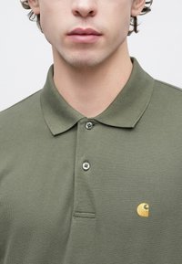 Πράσινο πουκάμισο πόλο σε χρώμα ελιάς, κατασκευασμένο από υφασμάτινη υφή, με διπλωμένο γιακά, δύο κουμπιά και ένα μικρό χρυσό λογότυπο στο αριστερό στήθος.