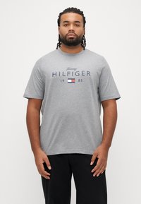 Szary bawełniany t-shirt z okrągłym dekoltem i krótkimi rękawami, z napisem "Tommy HILFIGER 1985" oraz logo w granatowo-czerwonych kolorach na przodzie.