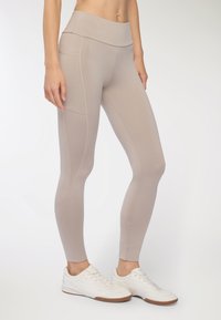 Beżowe legginsy z wysokim stanem o gładkiej fakturze, z bocznymi szwami i dopasowanym krojem. Połączone z białymi butami sportowymi.
