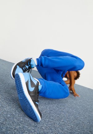 Personne portant un pantalon de survêtement bleu et des baskets noires et bleues effectue un mouvement de breakdance sur un sol recouvert de moquette grise contre un mur blanc.