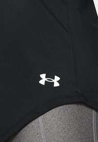 Gros plan sur un tissu sportif noir avec un logo blanc Under Armour au-dessus d’un tissu gris texturé avec des coutures.