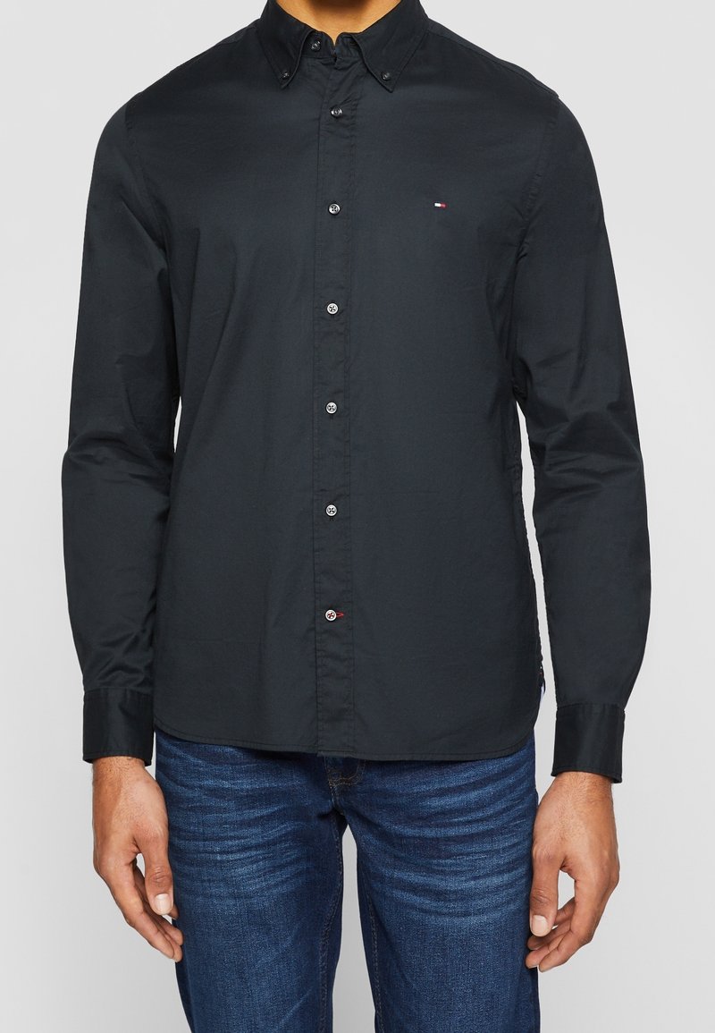 Musta napitettava kauluspaita, jossa on kaulus, pitkät hihat ja hienovarainen logoyksityiskohta. Paidassa on sileä pinta ja metalliset napit sulkemista varten.