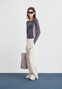 Μωβ μακρυμάνικη μπλούζα με ανοιχτό ροζ πλαϊνά πάνελ, λευκό λογότυπο Nike στο στήθος. Φαρδιά μπεζ φόρμα με μπλε λογότυπο Nike. Μπεζ τσάντα.