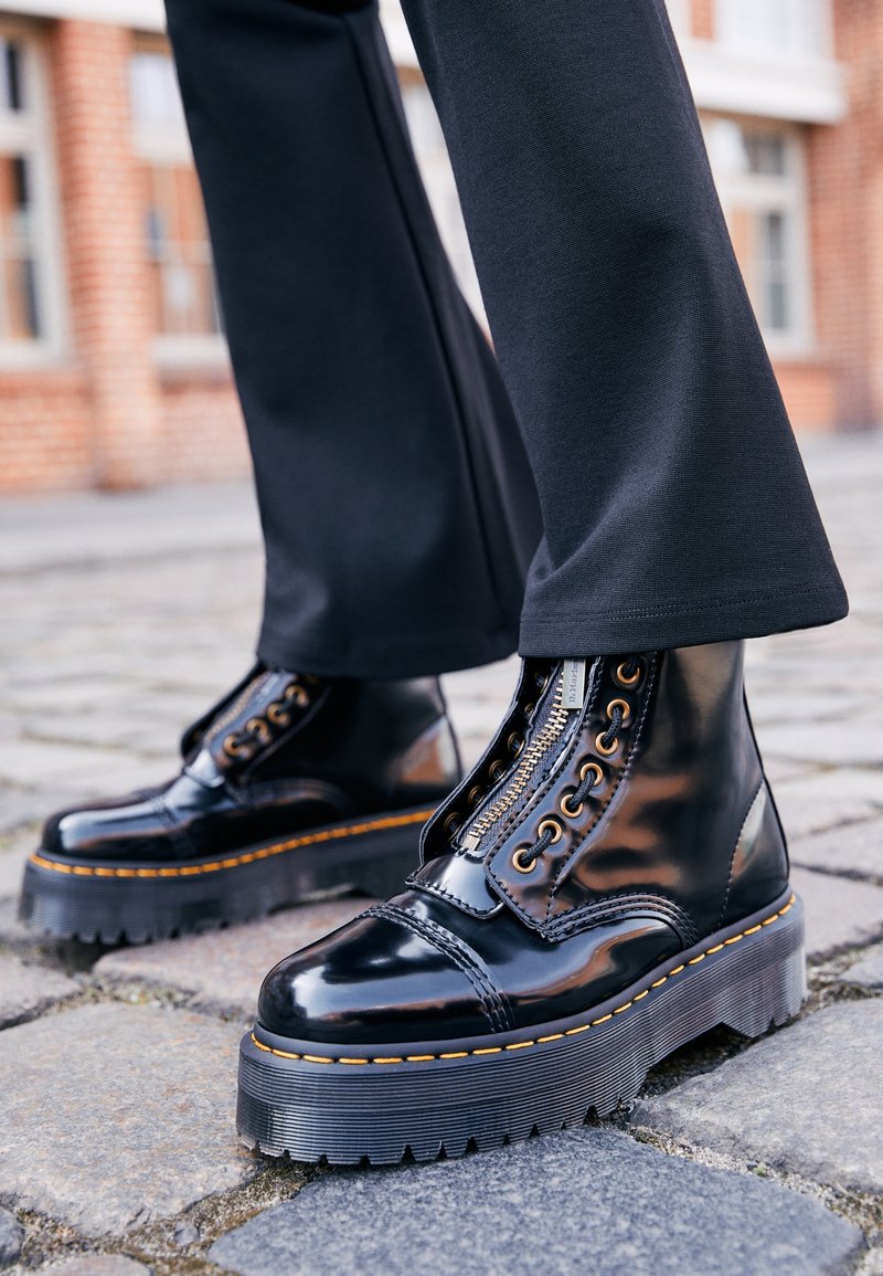 Bottes plateformes noires brillantes avec fermeture éclair frontale et coutures jaunes, portées avec un pantalon noir dans une rue pavée.