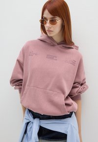 Felpa rosa con tasca frontale, vestibilità oversize e dettagli testuali. Abbinata a pantaloni shorts in denim e una camicia a righe blu chiaro legata in vita.