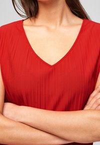 Czerwona, bezrękawnikowa bluzka z dekoltem w kształcie litery V, z teksturowanymi fałdami i gładkim materiałem. Dekolt jest wykończony, a ramiączka są lekko drapowane.