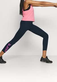 Kvinna i rosa linne och marinblå leggings som gör ett utfall framåt med armarna sträckta, iklädd svarta sportskor på en enfärgad bakgrund.