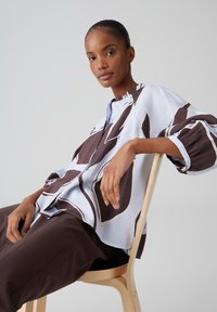 Frau sitzt seitlich auf einem hölzernen Stuhl, trägt ein gemustertes, übergroßes Hemd und dunkle Hosen vor einem schlichten hellen Hintergrund.