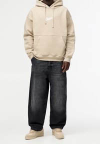 Beige Kapuzenpullover mit einer Fronttasche und weißem Logo, kombiniert mit lockeren, dunkelgrauen Jeans und beigen Turnschuhen.