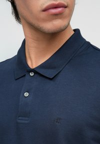 Tengerészkék póló, textúrált anyagból. Három gombbal és hímzett logóval a bal mellen. Klasszikus gallérterv.