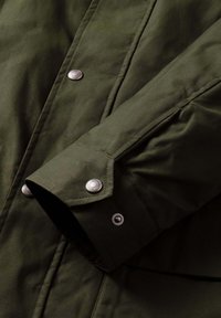 Chaqueta verde oscuro con puño abotonado, con herrajes plateados y textura suave. El diseño incluye detalles de costuras visibles.