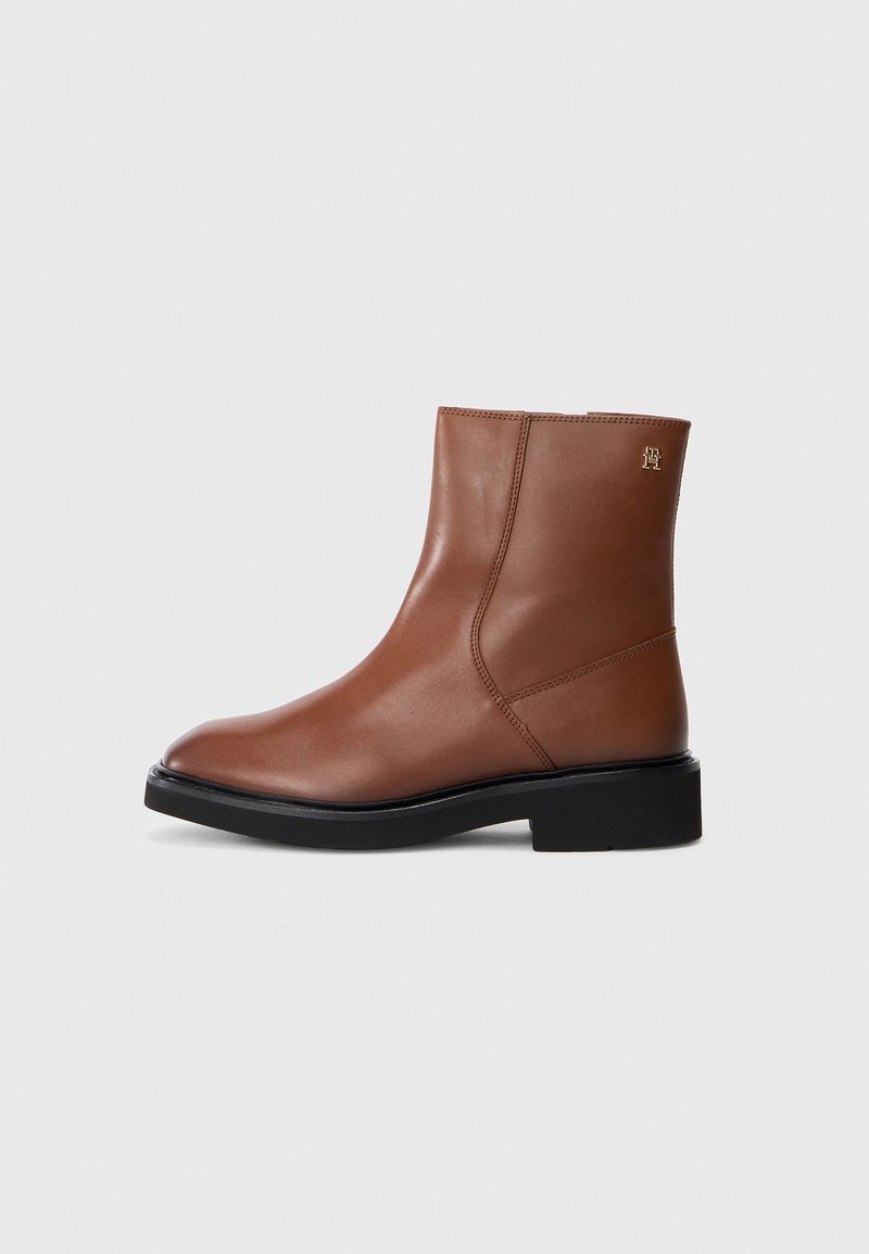 Bottine en cuir marron à la cheville avec semelle noire et talon bas, détails de coutures latérales et petit logo doré près du bord supérieur.
