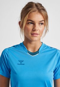 Blå sports trøje med korte ærmer, en v-hals, og kontrasterende hvid trim. Har et logo på venstre side af brystet.