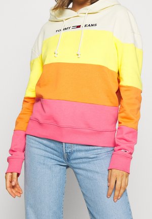 Person trägt einen mehrfarbigen Hoodie mit gelben, orangefarbenen und pinken Streifen, hellblaue Jeans und das "Tommy Jeans"-Logo auf der Brust.