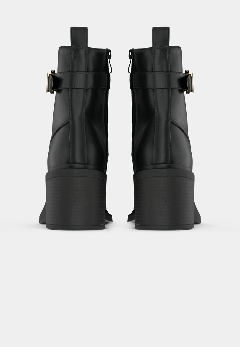 Paire de bottines noires à talons carrés, fermeture éclair à l'arrière et bride avec détail de boucle dorée, vues de dos sur un fond clair.