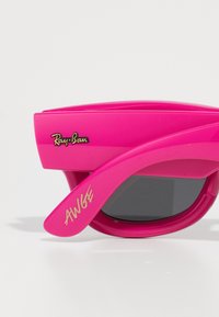 Ružičaste plastične sunčane naočale s crnim lećama. Značajke uključuju istaknuti Ray-Ban logo i stilizirani tekst "AWGE" na dršci.