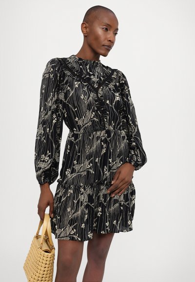 Vestido negro con flores, de mangas largas, con escote volado y dobladillo en capas. Presenta patrones florales en blanco y una cintura ajustada. Sosteniendo un bolso de mimbre amarillo.