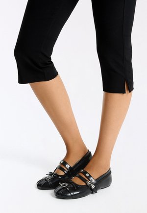 Jambes portant un pantalon noir court et des chaussures Mary Jane en cuir noir avec doubles brides et boucles argentées sur fond blanc.