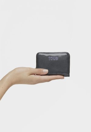 Mano sosteniendo una pequeña cartera con cremallera TOUS negra brillante frente a un fondo blanco liso.