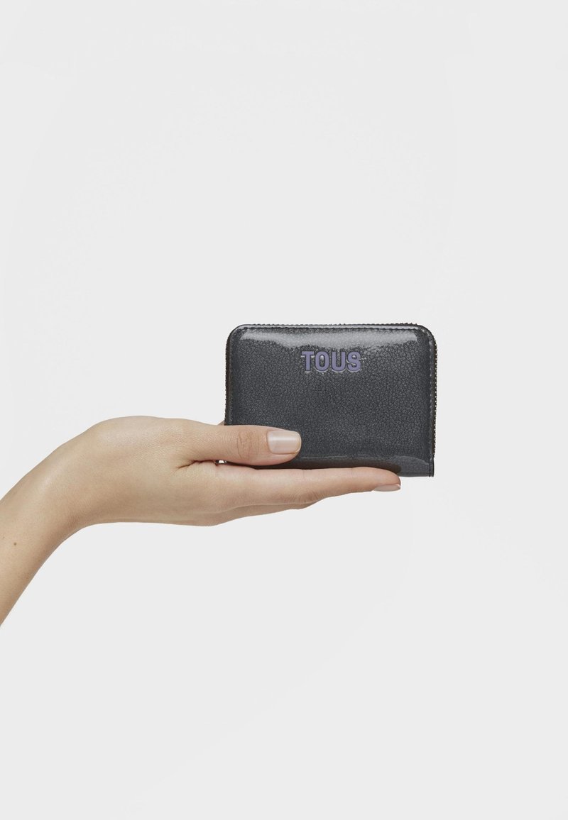 Ręka trzyma mały, błyszczący czarny portfel TOUS z suwakiem na tle gładkiego białego tła.
