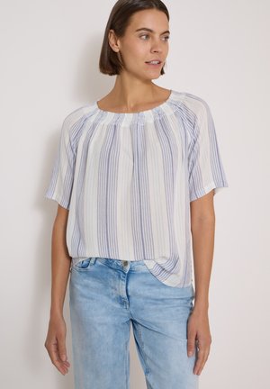 Frau trägt eine lockere weiße Bluse mit vertikalen blauen Streifen und hellblaue Jeans, steht vor einem einfarbigen hellen Hintergrund.