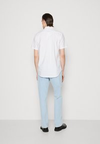 Mężczyzna stoi twarzą do białej ściany, ubrany w białą koszulę z krótkim rękawem, jasnoniebieskie dżinsy i czarne buty.