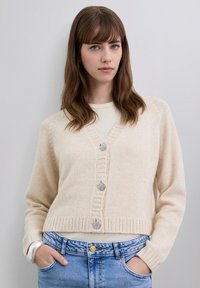 Mujer joven con cabello marrón ondulado, lleva un cárdigan corto color crema con botones decorativos y vaqueros de talle alto en azul claro.