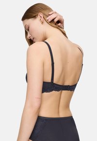 Žena koja sređuje svoju dugu kosu, odjevena u crni čipkasti grudnjak i odgovarajuće gaćice visokog struka, okrenuta leđima prema jednostavnoj pozadini.