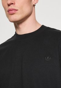 Tricou negru din bumbac, cu guler rotund și un mic logo brodat în negru pe pieptul stâng. Textură netedă și design ajustat.