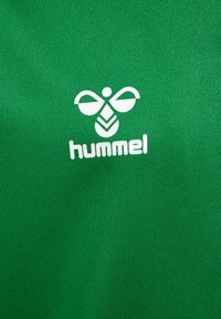 Grøn tekstilskjorte med et hvidt logo med abstrakte former og brandnavnet "hummel" nederst, på et glat, vævet stof.