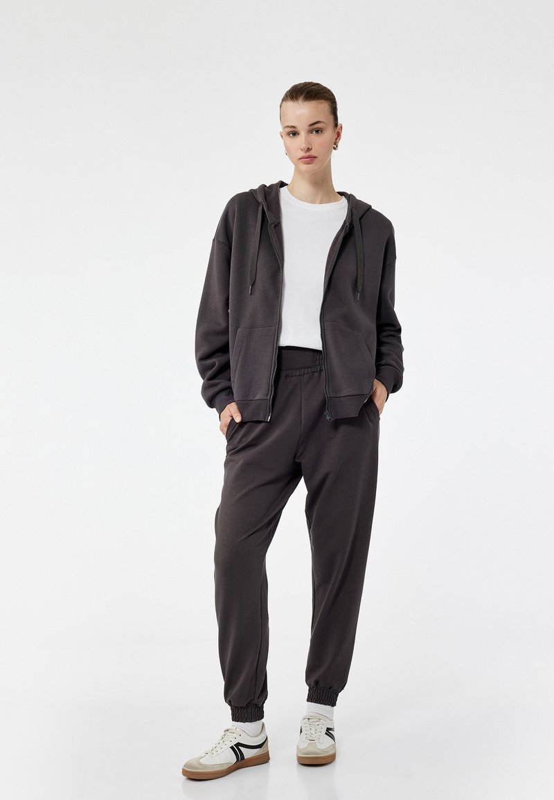 Donkergrijze zip-up hoodie met twee voorkeuzes, gecombineerd met bijpassende joggers met elastische boorden. Gekleed met een wit t-shirt en beige sneakers.