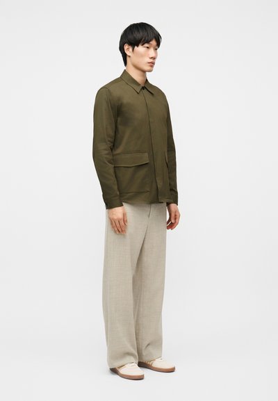 Νεαρός άνδρας όρθιος, φορώντας ένα χακί πράσινο μπουφάν με τσέπες με καπάκι, μπεζ παντελόνι με φαρδιά μπατζάκια και ελαφρώς μπεζ παπούτσια.