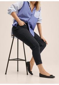 Camisa a rayas azul y blanca superpuesta sobre unos jeans skinny negros, combinada con unas bailarinas negras con un acento de lazada. Sentada en un taburete.
