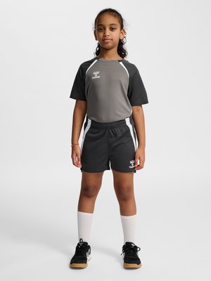 Ung pige står, iført sort og gråt sportstøj med hvide striber, hvide knæstrømper og sorte sneakers.