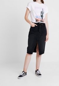Falda de mezclilla negra con abertura frontal, dobladillo deshilachado, combinada con una camiseta gráfica blanca que presenta un diseño de cactus y mangas enrolladas.