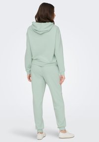 Zelena majica z dolgimi rokavi in trenirke iz mehkega blaga. Majica ima kapuco; trenirke imajo elastične manšete in sproščen kroj.