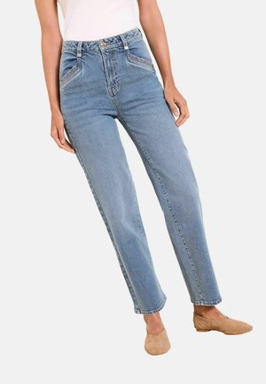 Woman wearing high-waist light blue jeans with slanted front pockets and tan suede flat shoes against a white background.