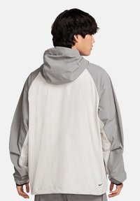 Nike-jack met capuchon, met een lichtgrijze romp en donkergrijze mouwen, gemaakt van een lichtgewicht, glad materiaal.