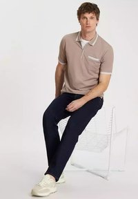 Beżowa koszulka polo z białymi detalami, krótkim rękawem i kieszonką na piersi. Ciemnoniebieskie dżinsy i jasne trampki dopełniają stylizację.