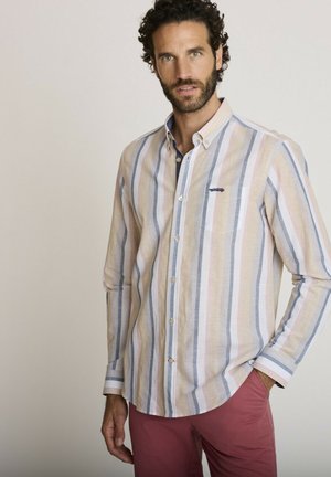 Homme portant une chemise à rayures pastel et un pantalon rouge, debout avec une main dans la poche sur un fond uni.