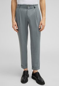 Pantaloni grigi chiari su misura con pieghe, dotati di chiusura con bottone e passanti per cintura. Cuffie arrotolate e texture liscia, abbinati a scarpe nere.