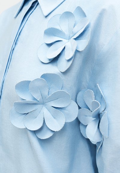 Cămașă bleu deschis cu flori mari, stratificate, din același material de culoare similară. Materialul are o textură netedă și design cu guler.