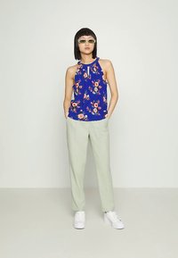 Blusa azul com padrão floral e detalhe gota no decote, combinada com calças verdes claras e ténis brancos. Design elegante, estilo casual.