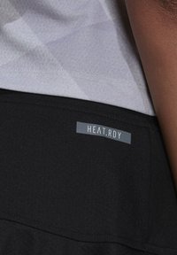 Camisa deportiva negra con una textura suave, que presenta una etiqueta gris con la inscripción "HEAT.RDY." y una sección superior de un gris claro con patrón.