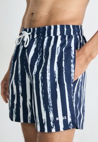 Badeshorts in Marineblau mit weißem Pinselstrichmuster. Elastischer Bund mit Kordelzug und Seitentaschen. Logo unten links.