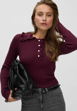 Femme portant un pull côtelé violet foncé avec col à volants et boutons, tenant un sac en cuir noir, avec un jean gris et des cheveux longs.